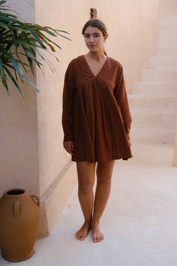 A woman poses on a patio wearing our Vintage Collar Dress, a joint design by Audrey E Leary and World of Crow. This dress, in deep chocolate brown, showcases a timeless collar and full sleeves, made from handwoven cotton for an elegant and eco-friendly appearance.