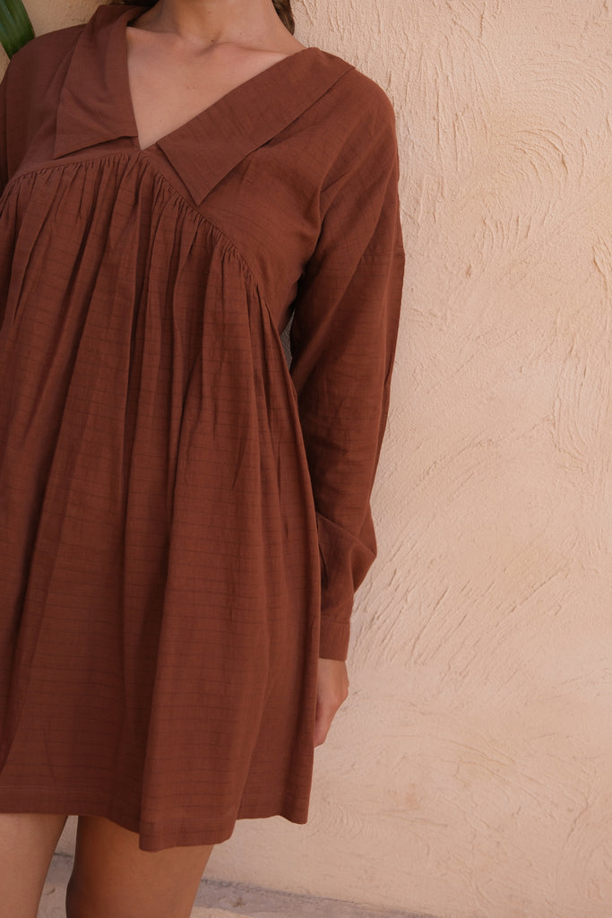 A woman poses on a patio wearing our Vintage Collar Dress, a joint design by Audrey E Leary and World of Crow. This dress, in deep chocolate brown, showcases a timeless collar and full sleeves, made from handwoven cotton for an elegant and eco-friendly appearance.